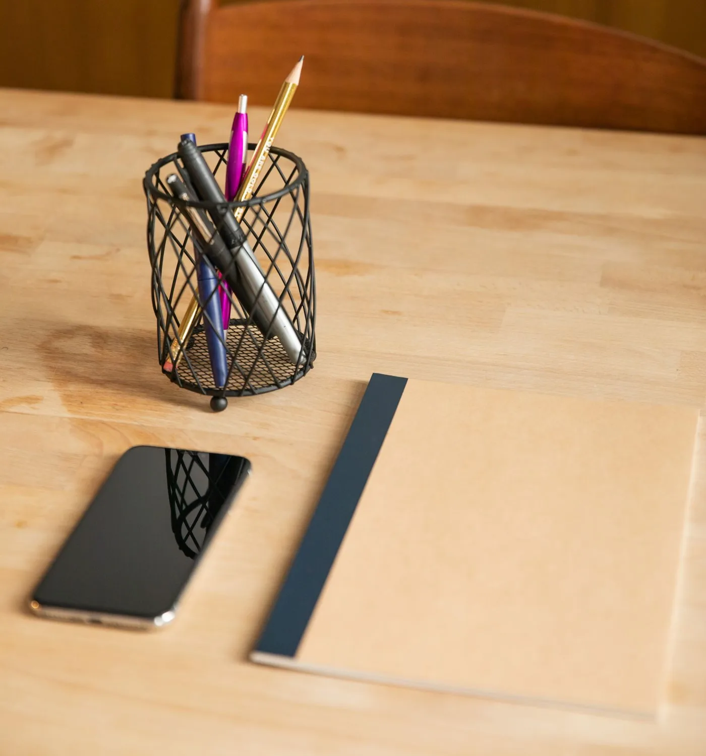 木の机に置かれた黒いスマートフォンと、ペンが挿さったワイヤー製のペン立て、閉じたノート。