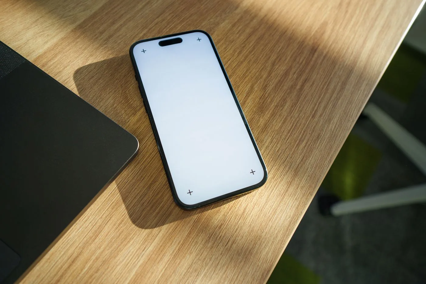 木目調のデスクに置かれた、画面が白紙のスマートフォン。手前左にノートパソコンの端が見える。