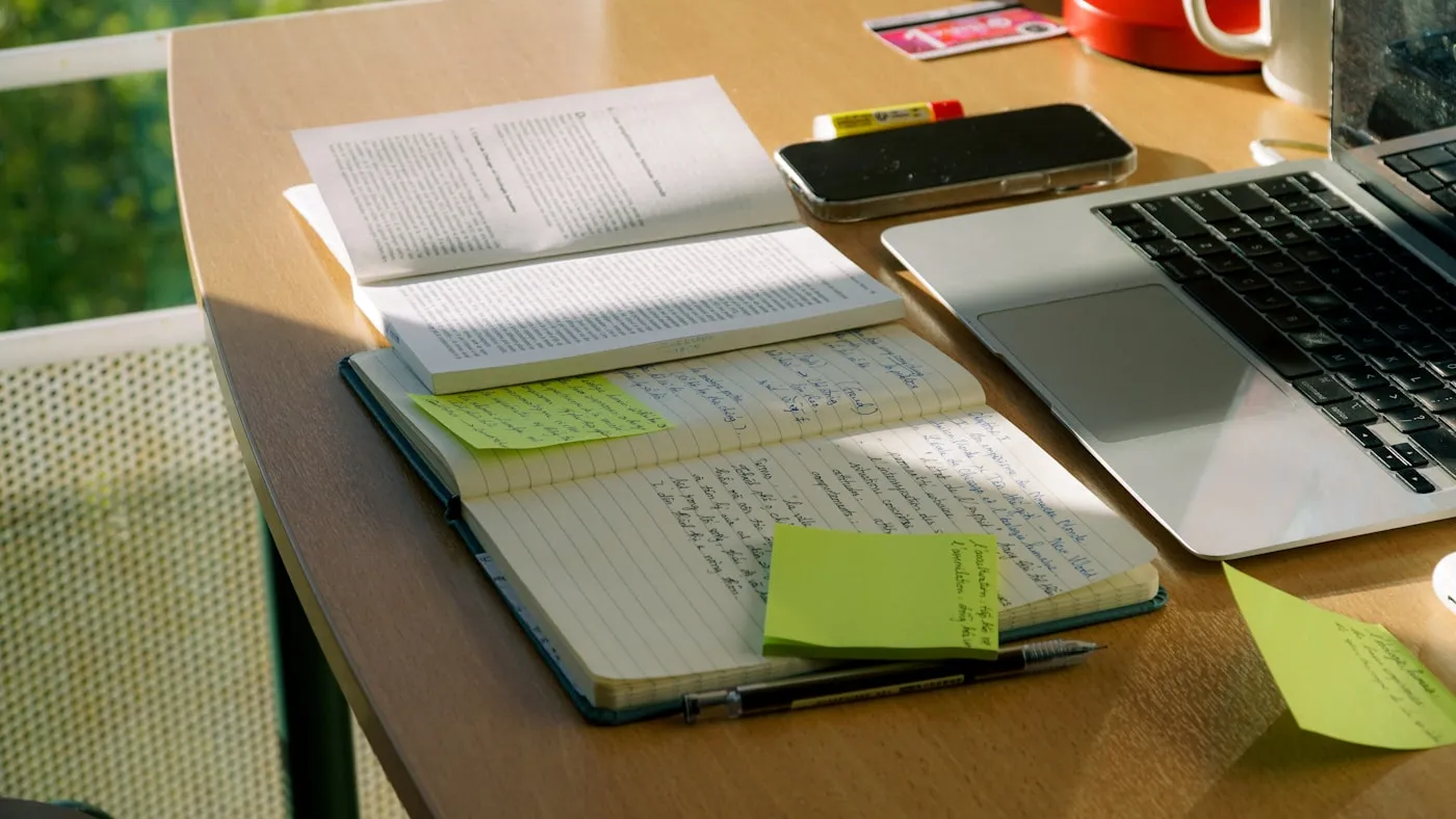 机の上に開いた本と書き込みのあるノート、付箋、ノートパソコンが並んでいる。