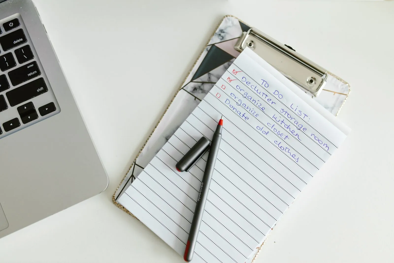赤いチェックの入った手書きのリストを挟んだクリップボードとペン。左にノートパソコンのキーボードが写っている。