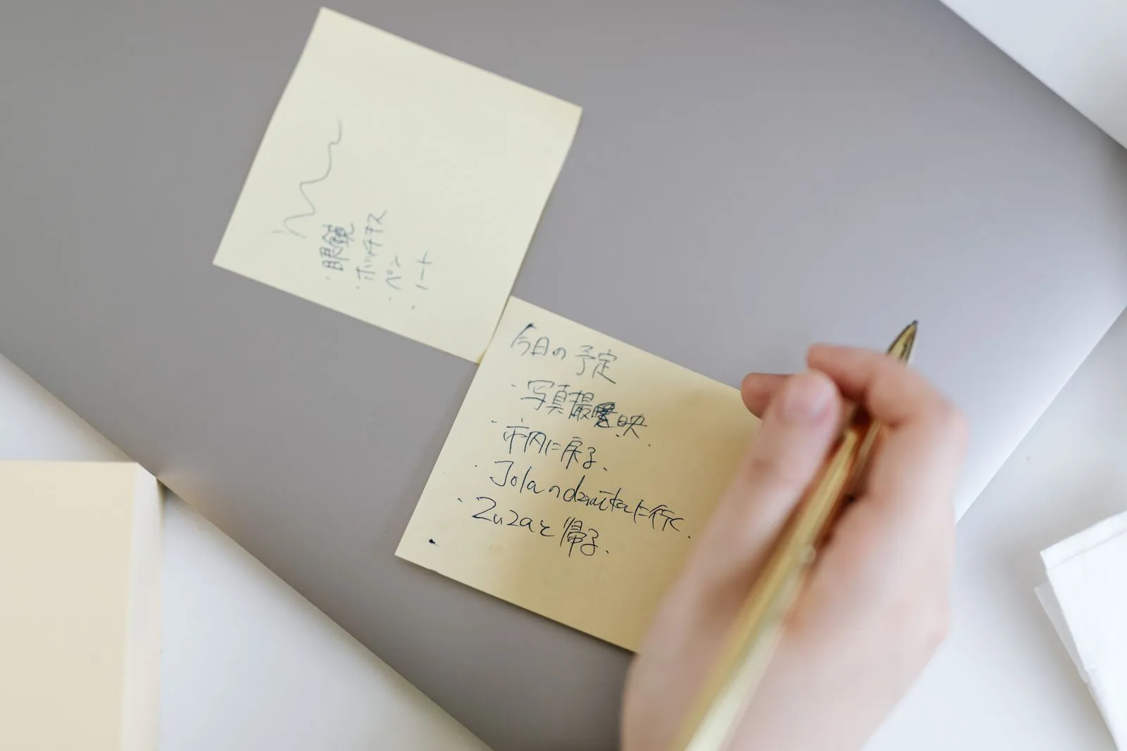 ノートパソコンの天板に貼られた2枚の黄色い付箋。手書きの項目が並び、右下にペンを持つ手が写っている。